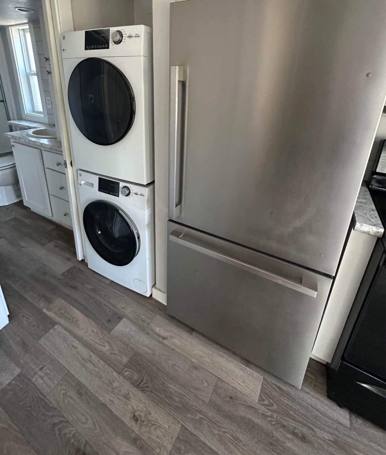 A compact laundry area shows a stacked washer and dryer next to a sleek stainless steel refrigerator. The space has light wood flooring and modern decor.