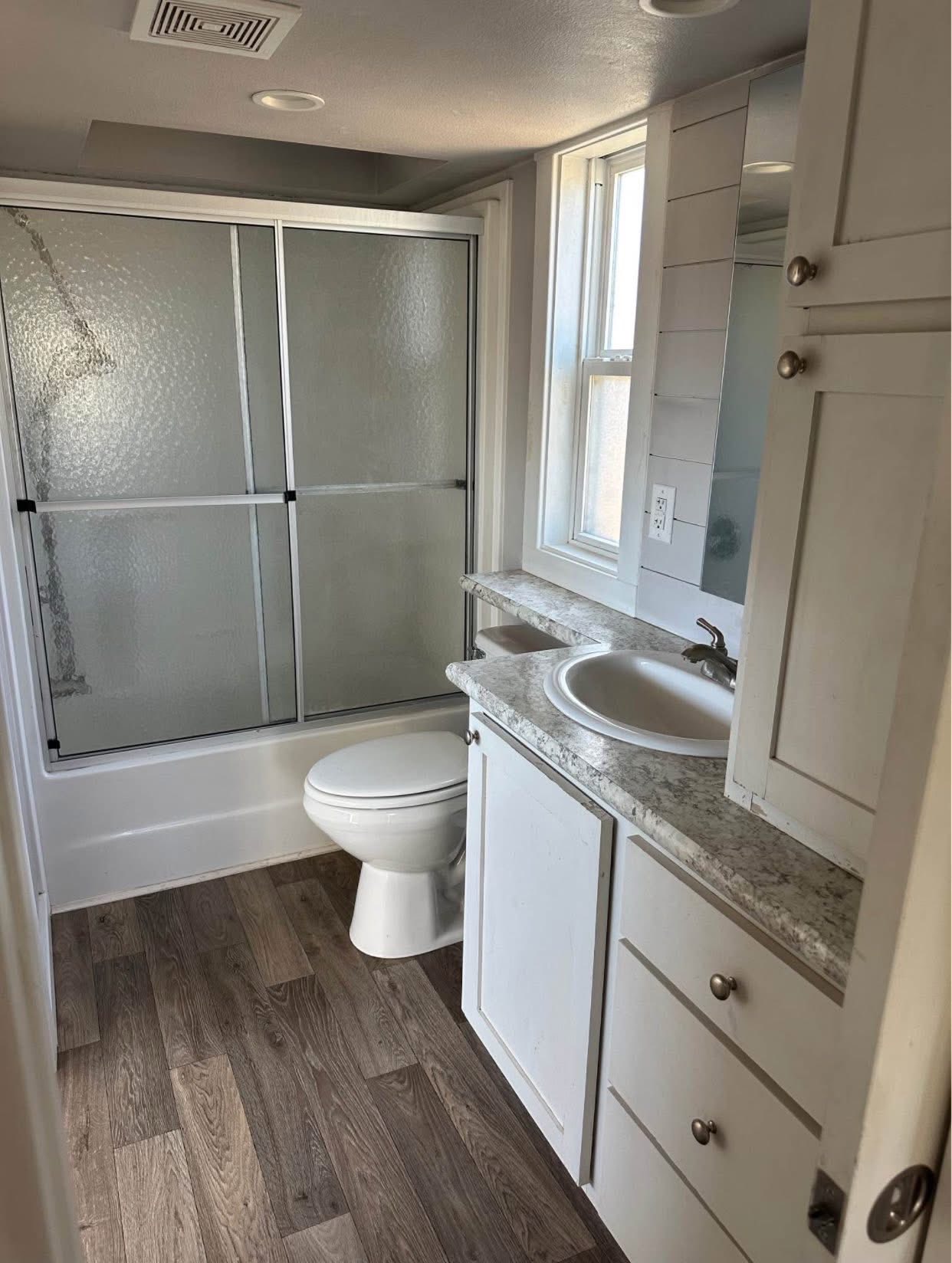 Small bathroom with wood-look flooring, a white toilet, and a shower with frosted glass doors. White vanity with a marble countertop and single round sink.