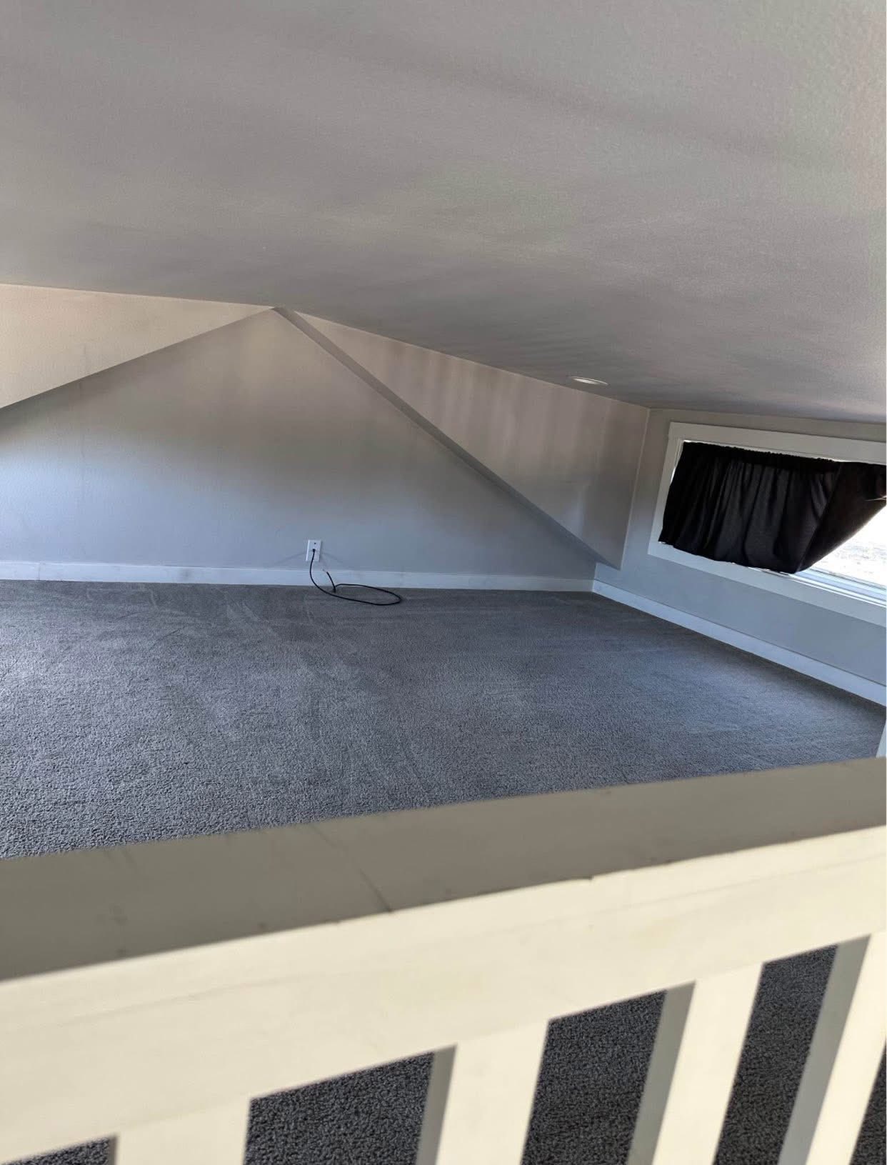 A small, carpeted attic space with sloped ceilings, a single window with dark curtains, and an electrical outlet visible on one wall. Cozy and minimal.