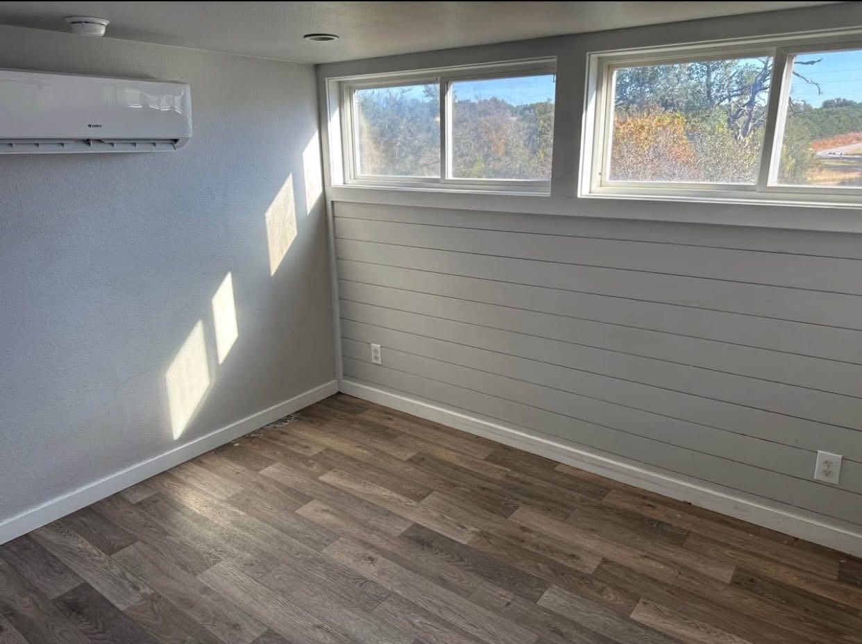 Small, sunlit room with light gray walls and wood flooring. Horizontal windows reveal a scenic view. Modern air conditioning unit mounted on the wall.