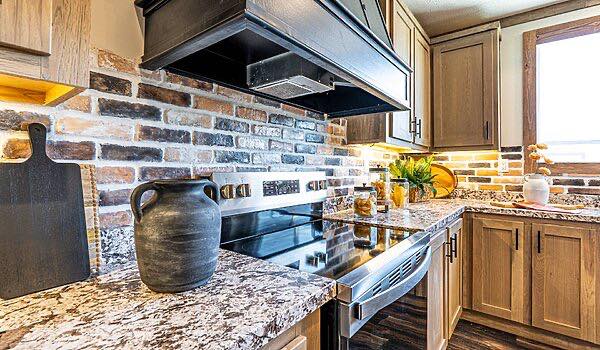Rustic kitchen featuring a granite countertop, black oven, and brick backsplash. Decor includes a black vase, cutting board, and potted plants. Warm lighting creates a cozy ambiance.