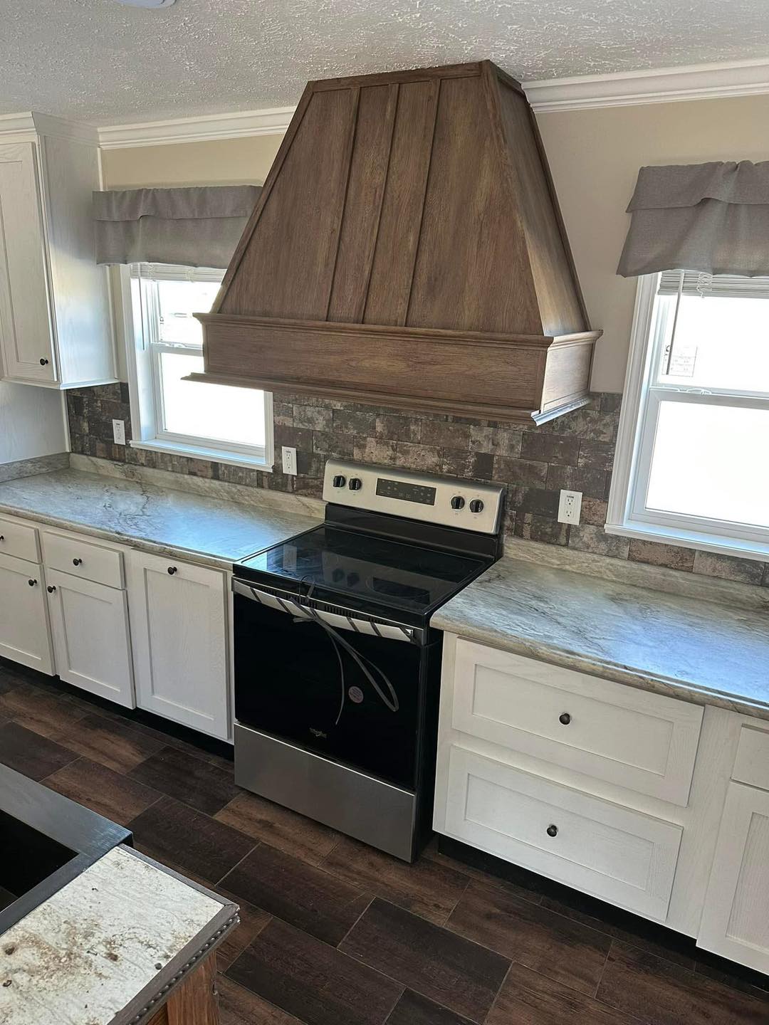 Modern kitchen with dark wood range hood, white cabinets, and stone backsplash. Stainless steel stove centered below, surrounded by marble countertops.