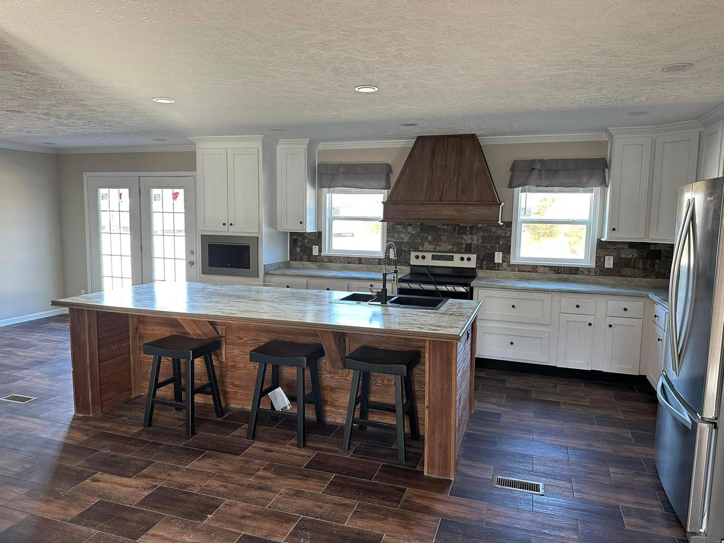 Modern kitchen with white cabinets, granite countertops, and a wooden island with three stools. Brick backsplash, stainless steel appliances, and dark wood floors. Natural light filters through two windows, creating a welcoming atmosphere.