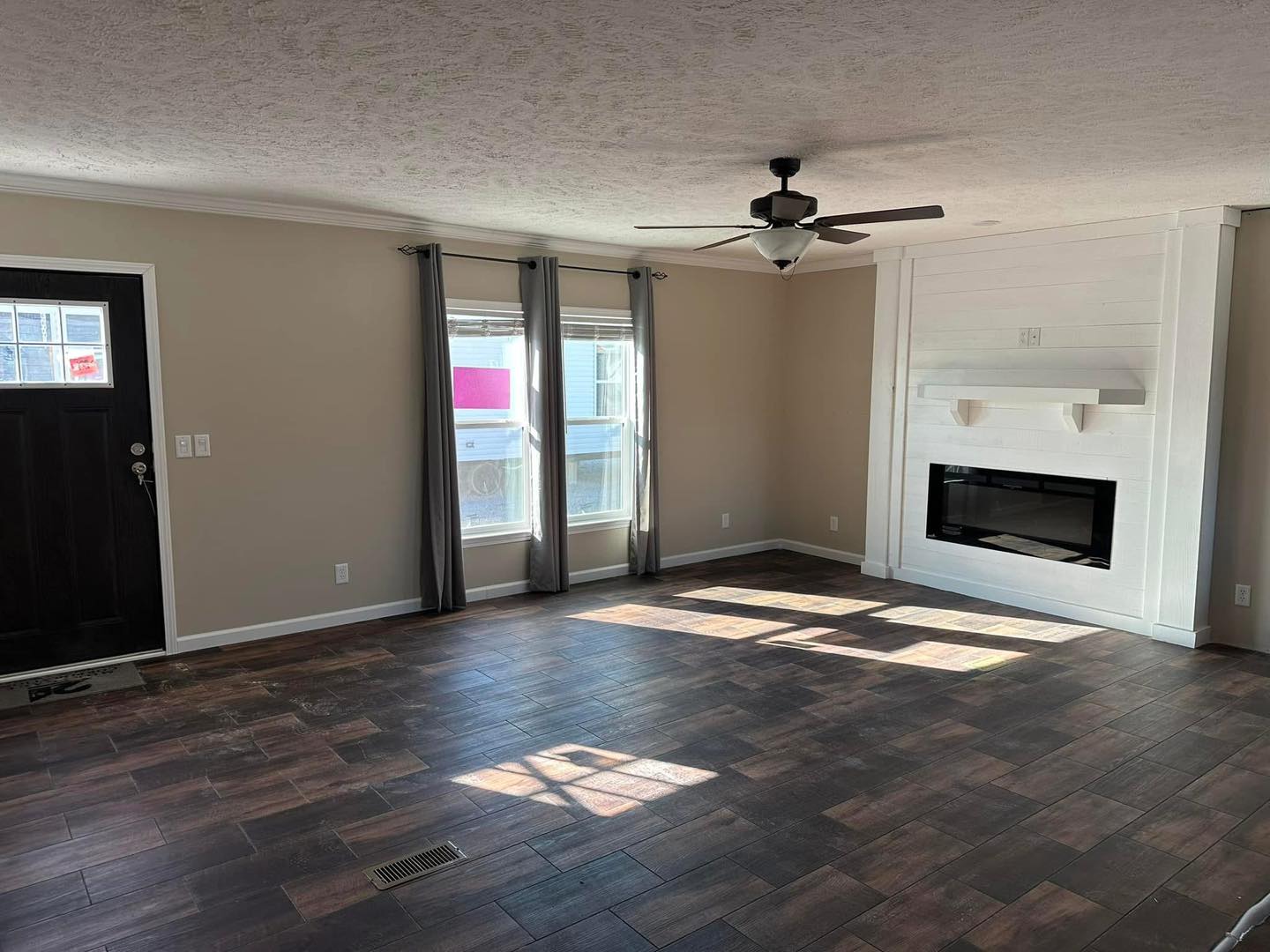 A spacious, empty living room with brown hardwood floors, beige walls, a dark wooden door, windows with gray curtains, a ceiling fan, and a white fireplace.