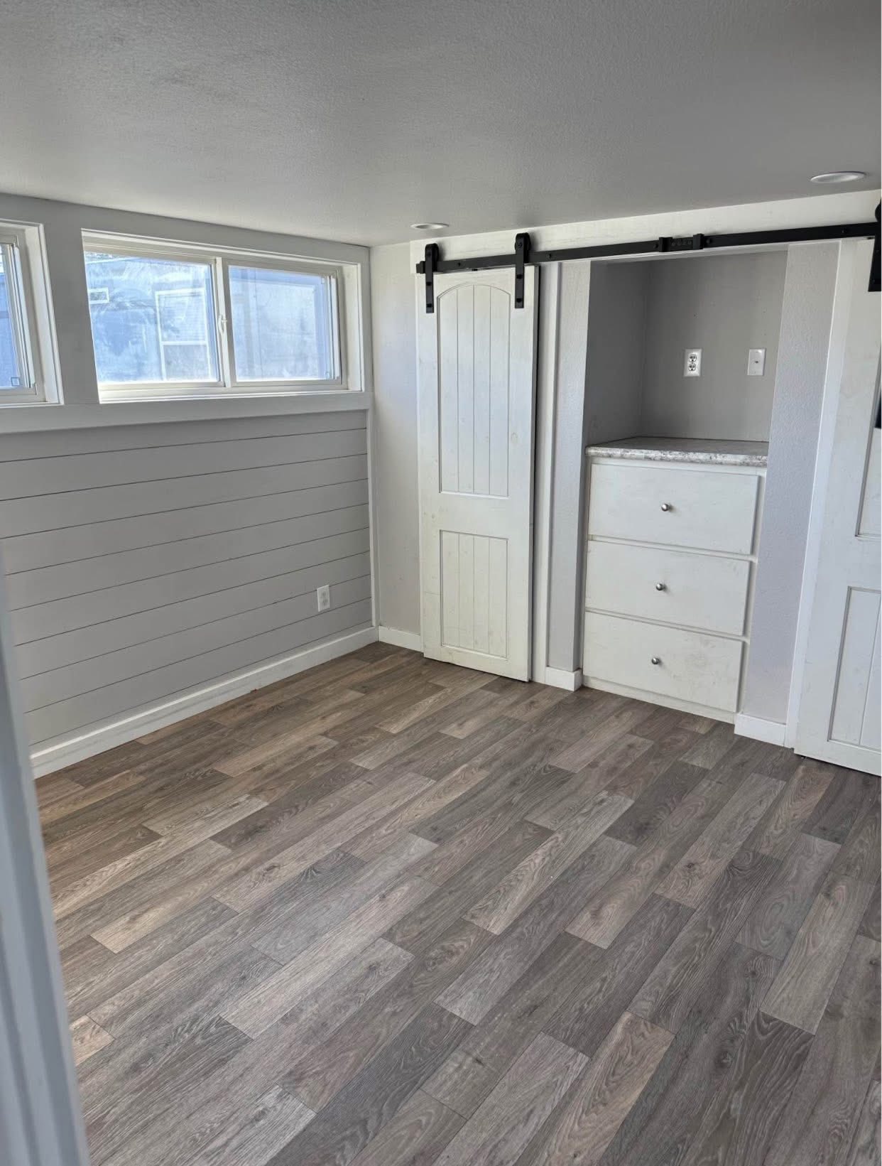 Cozy room with light gray paneled walls and wood-look flooring. It features large windows for natural light, a white sliding barn door, and built-in drawers.