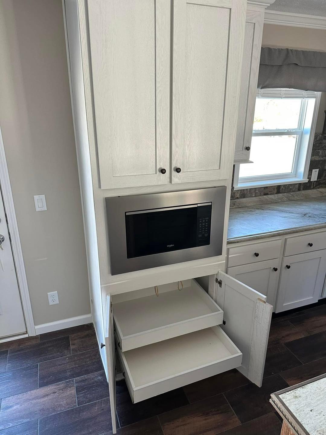 Tall kitchen cabinet with a built-in microwave and open storage drawers. Sunlight from a nearby window illuminates the sleek, modern design.