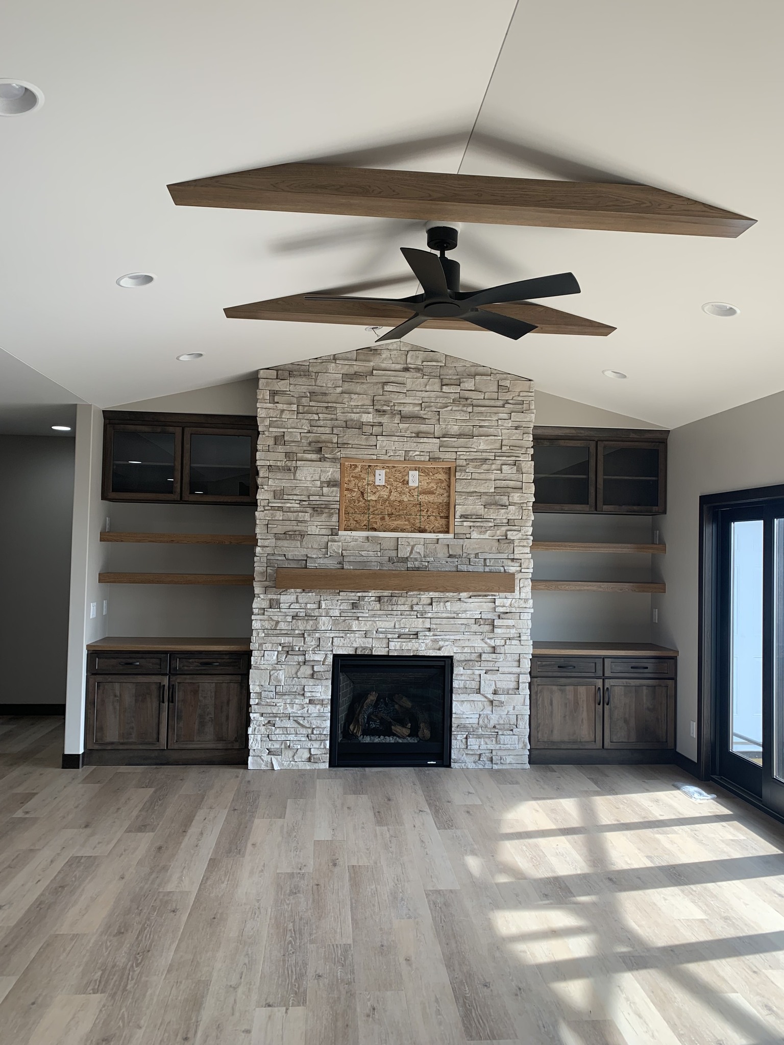 Spacious living room with light wood floors, a stone fireplace, and dark wood cabinets. A modern ceiling fan and natural light create a warm atmosphere.