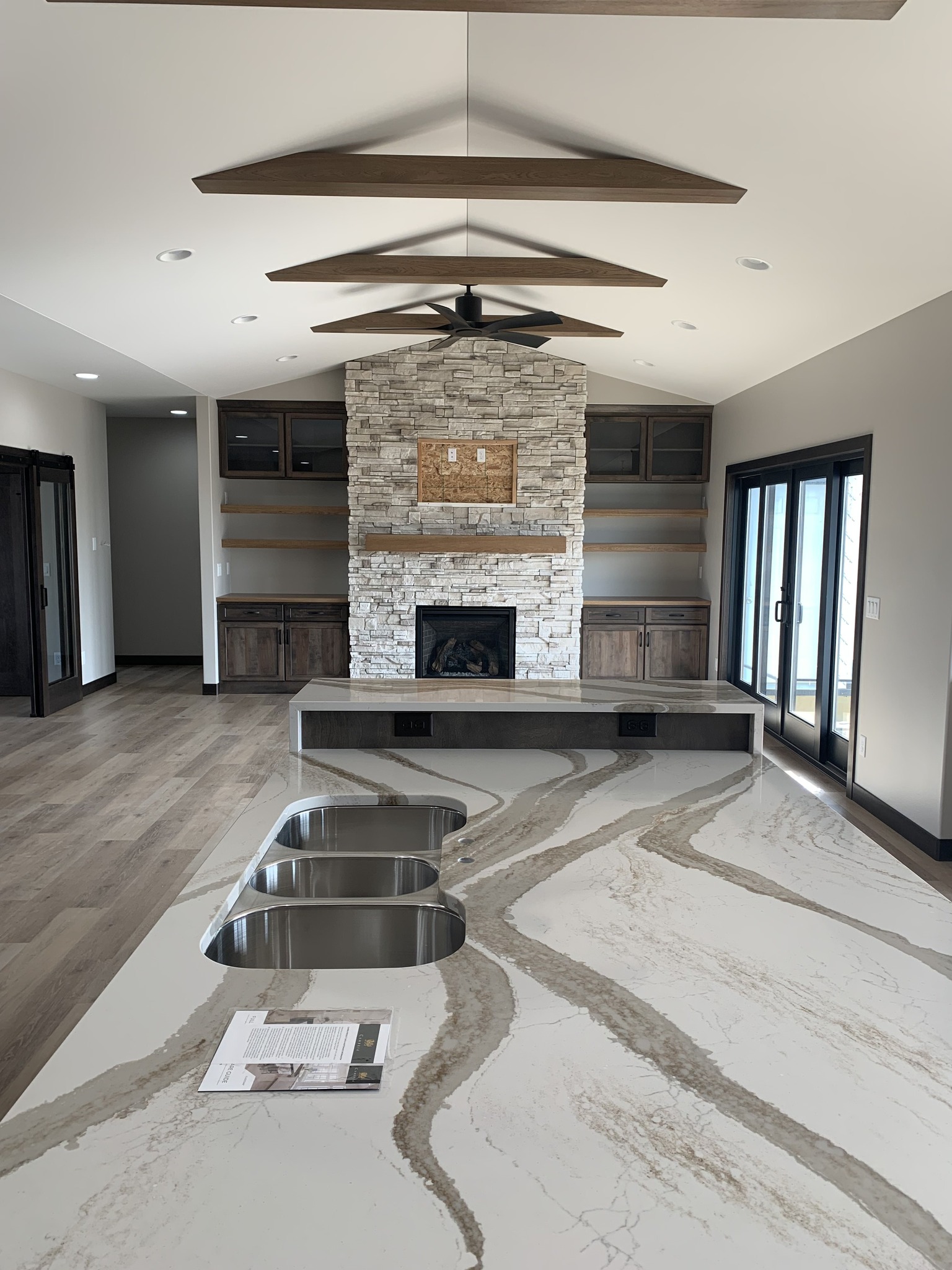 Modern kitchen and living area with a large marble island featuring two sinks. A stone fireplace is centered on a wall with wooden cabinets and shelves. The space is bright with natural light from large sliding doors.
