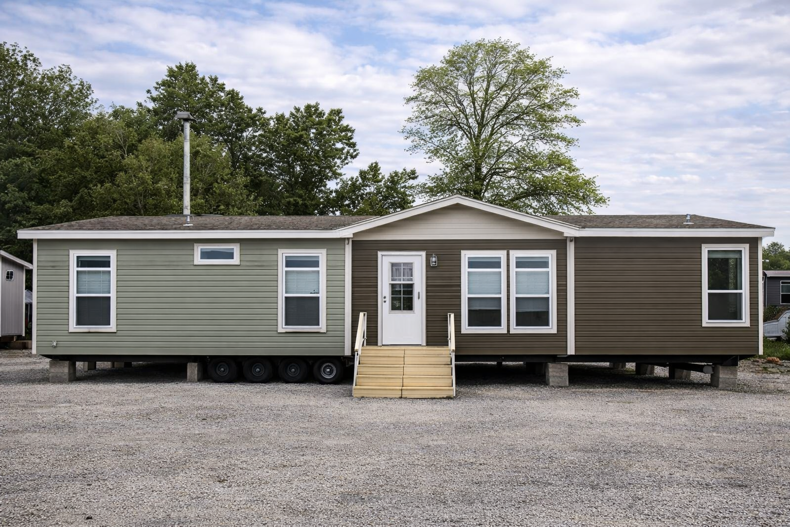 A modular home on a gravel lot, featuring a dual-toned exterior of green and brown siding, with a white door and a small wooden ramp leading to the entrance.