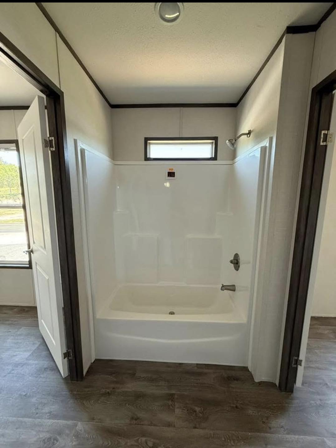 A modern bathroom with a white bathtub and shower is framed by two open doors. It features gray wood flooring and a small, high window allowing natural light.