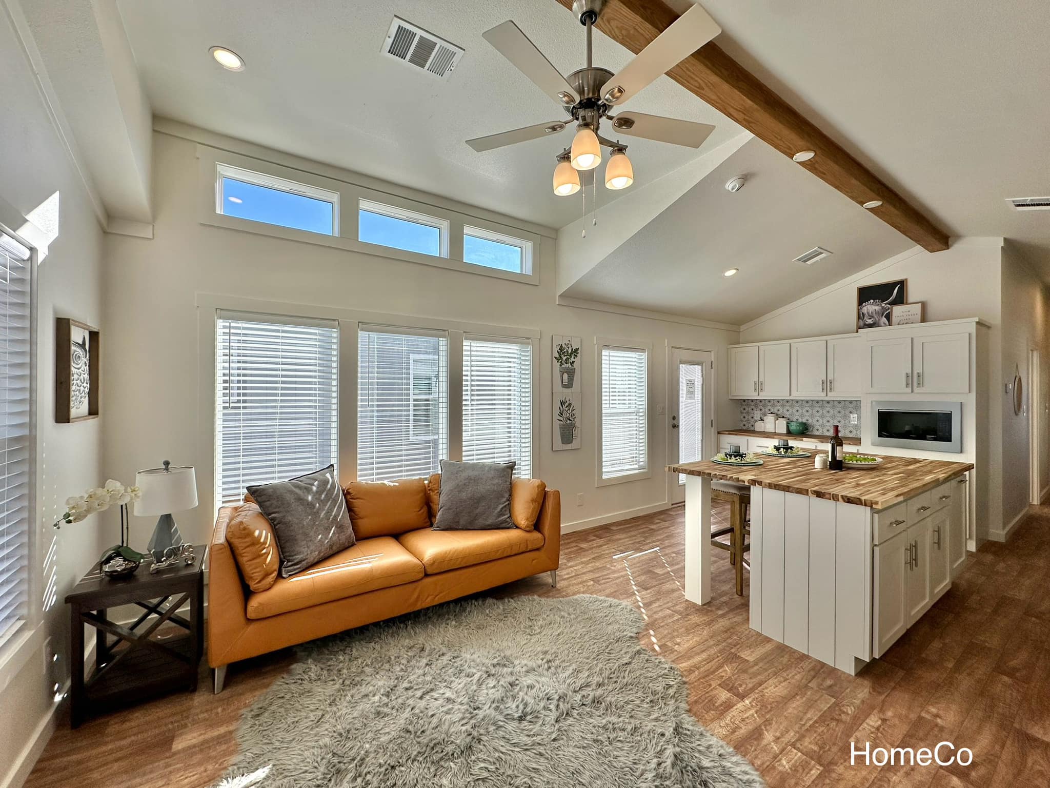 A cozy, sunlit living area features a mustard sofa with gray cushions, a plush rug, and a modern kitchen with a wooden island, creating a warm, inviting ambiance.