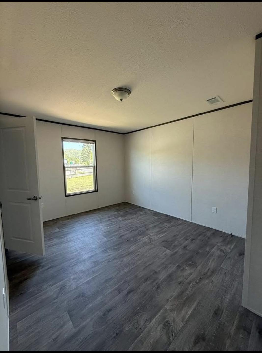 Empty room with light gray walls and dark wood flooring. A single window lets in daylight, and a ceiling light fixture hangs centrally. Minimal and serene.
