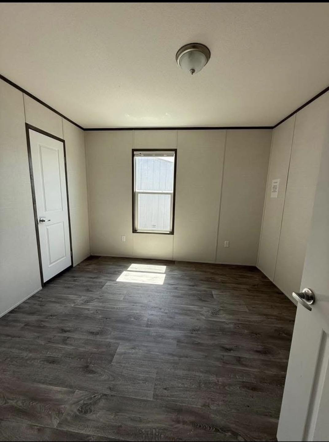 Empty room with beige walls and dark wood flooring. A window allows sunlight to spill onto the floor. There is a white door on the left wall.