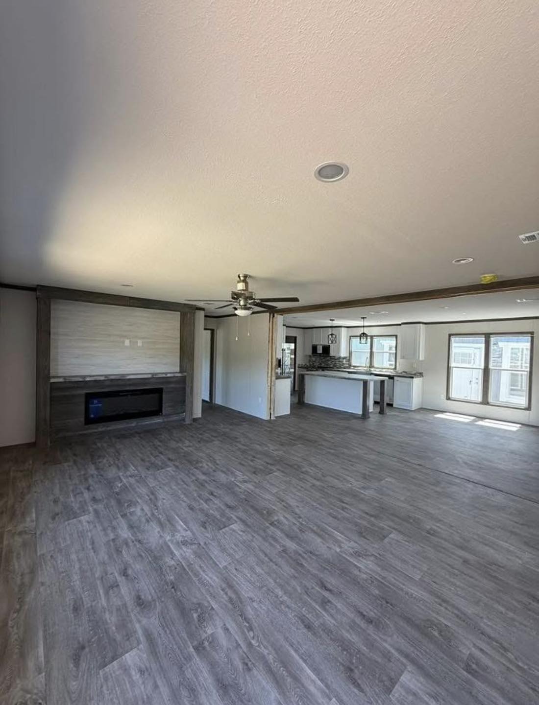Spacious open-concept living area with gray wood flooring, a ceiling fan, and a modern fireplace. Bright kitchen in background with large windows.