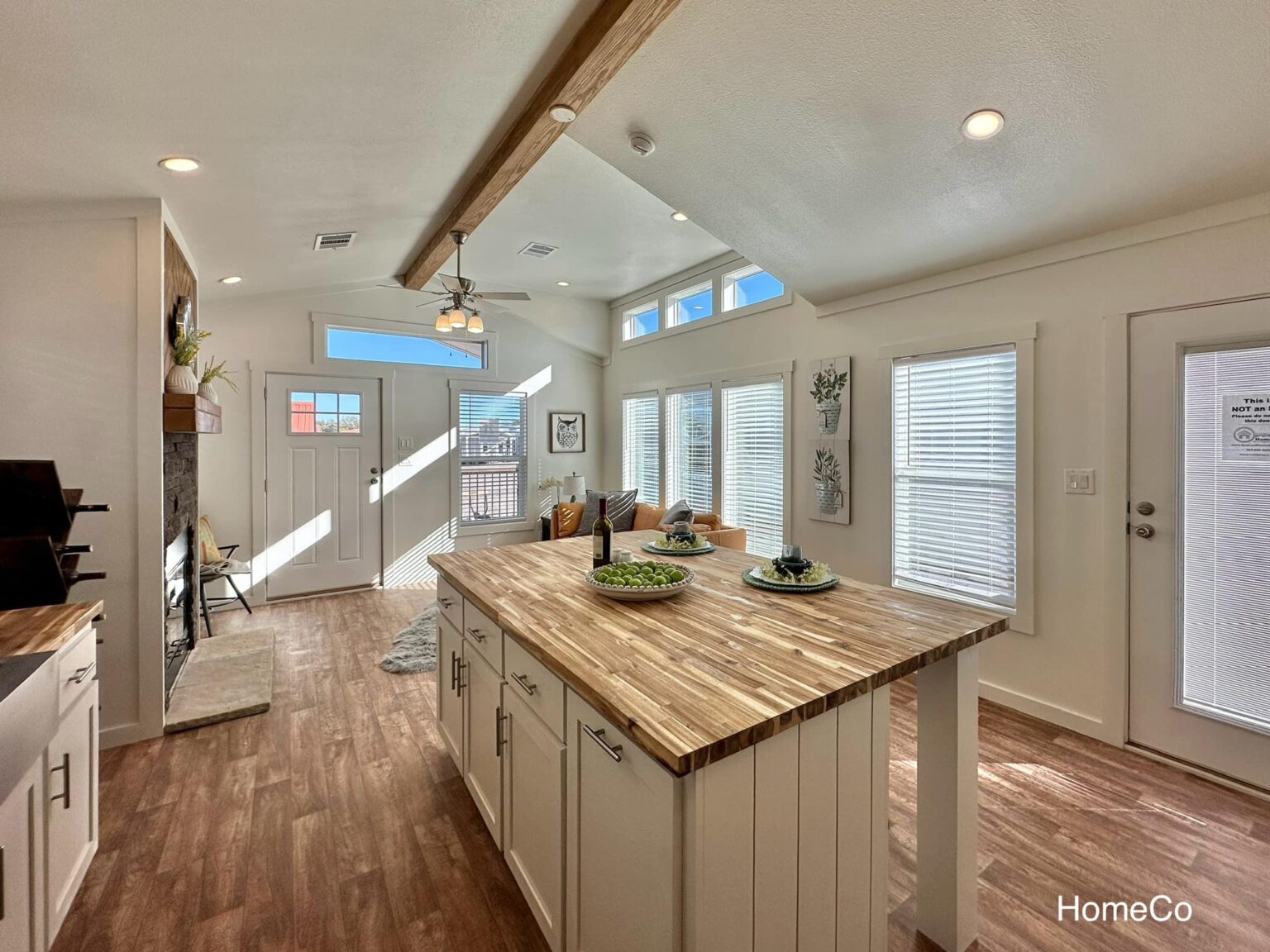Bright, modern kitchen with wood flooring, a spacious island, and large windows letting in sunlight. Cozy living area features a stone fireplace.