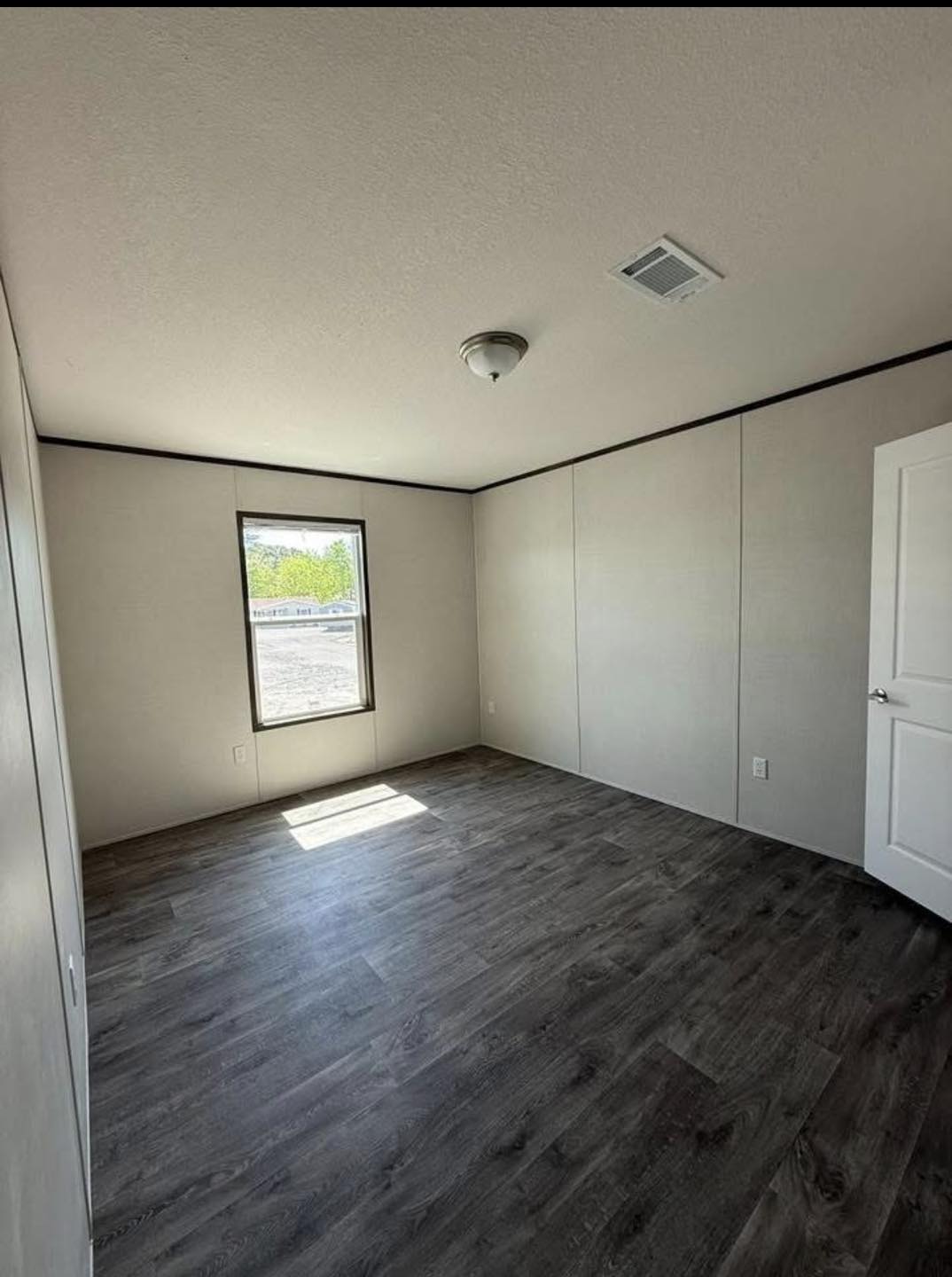 An empty room with light gray walls and dark hardwood floors. A single window lets in natural light. The door is open, and a ceiling light fixture is visible.