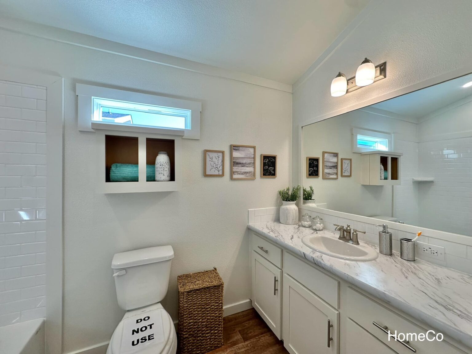 Bright, modern bathroom with a marble countertop, white cabinets, large mirror, wall art, decorative shelves, and a wicker basket beside the toilet.