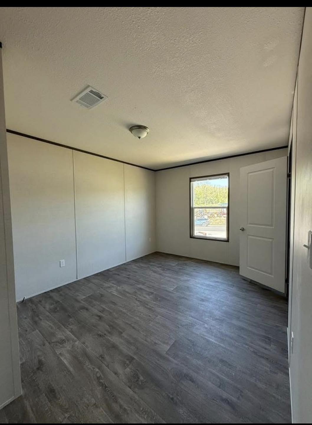 Empty room with light gray walls and dark wood laminate flooring. A window lets in natural light, and there's a white door slightly ajar.