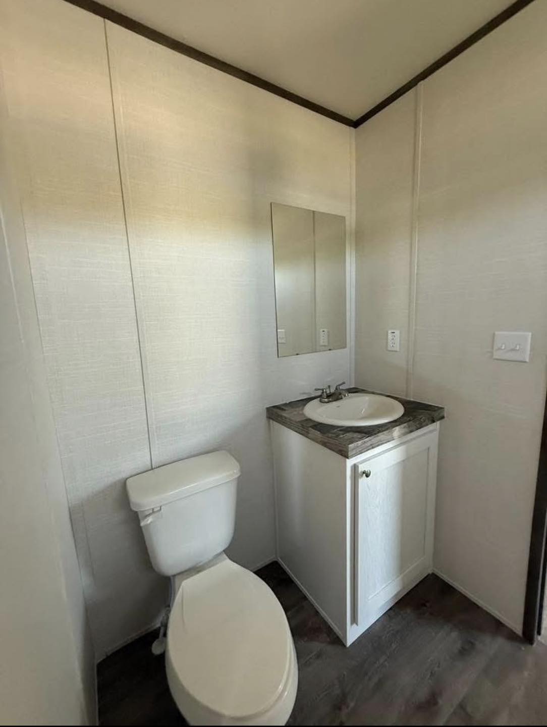 A small bathroom with light beige walls features a white toilet and a sink with a marbled countertop. Above the sink is a rectangular mirror. The room feels minimal and clean.