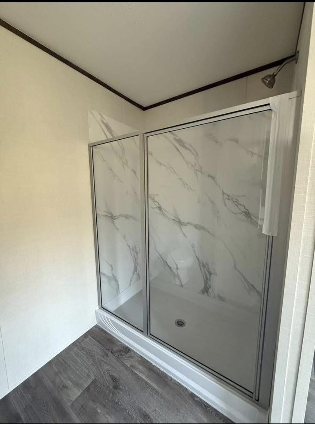 A modern bathroom shower with a glass door features a marble-patterned wall and chrome fixtures. The floor is dark wood, creating a clean, elegant look.