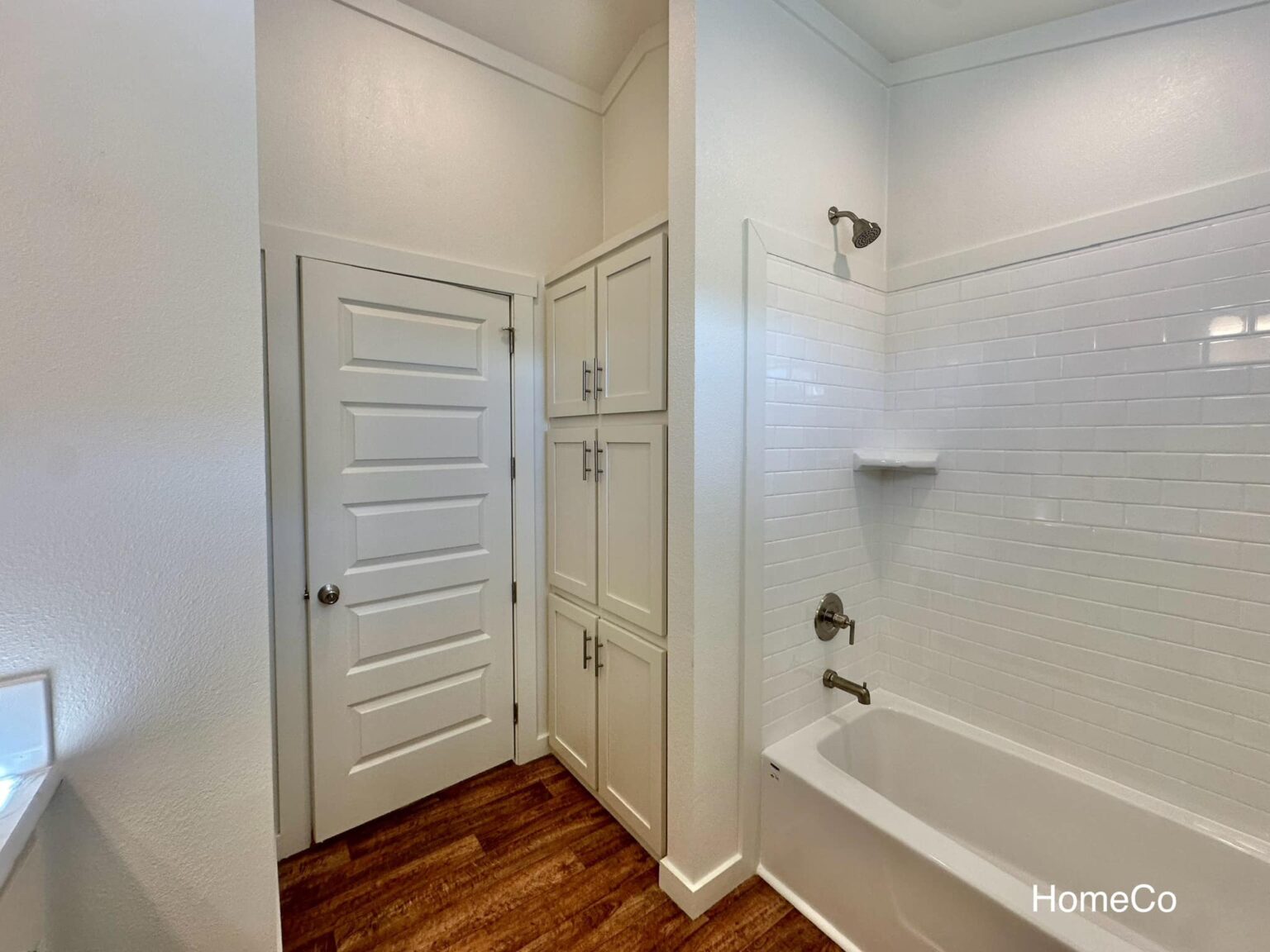 Bright bathroom with white walls, featuring a white bathtub and tiled shower on the right. On the left, there's a white door and storage cabinets. Dark wood floor.