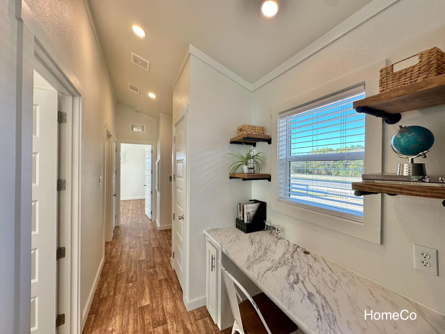 Bright hallway with wood flooring and white walls. A small desk with a marble top is on the right, flanked by shelves with decor. Warm, inviting ambiance.