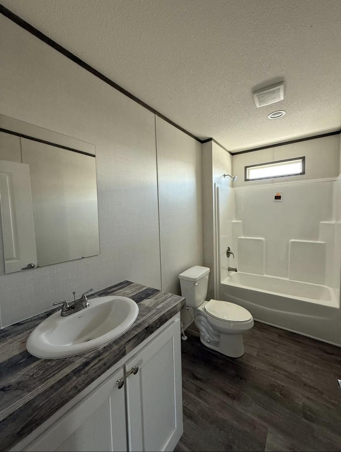 Modern bathroom with wood-look flooring, white vanity sink, mirror, and a toilet. A small window above a bathtub shower provides natural light.