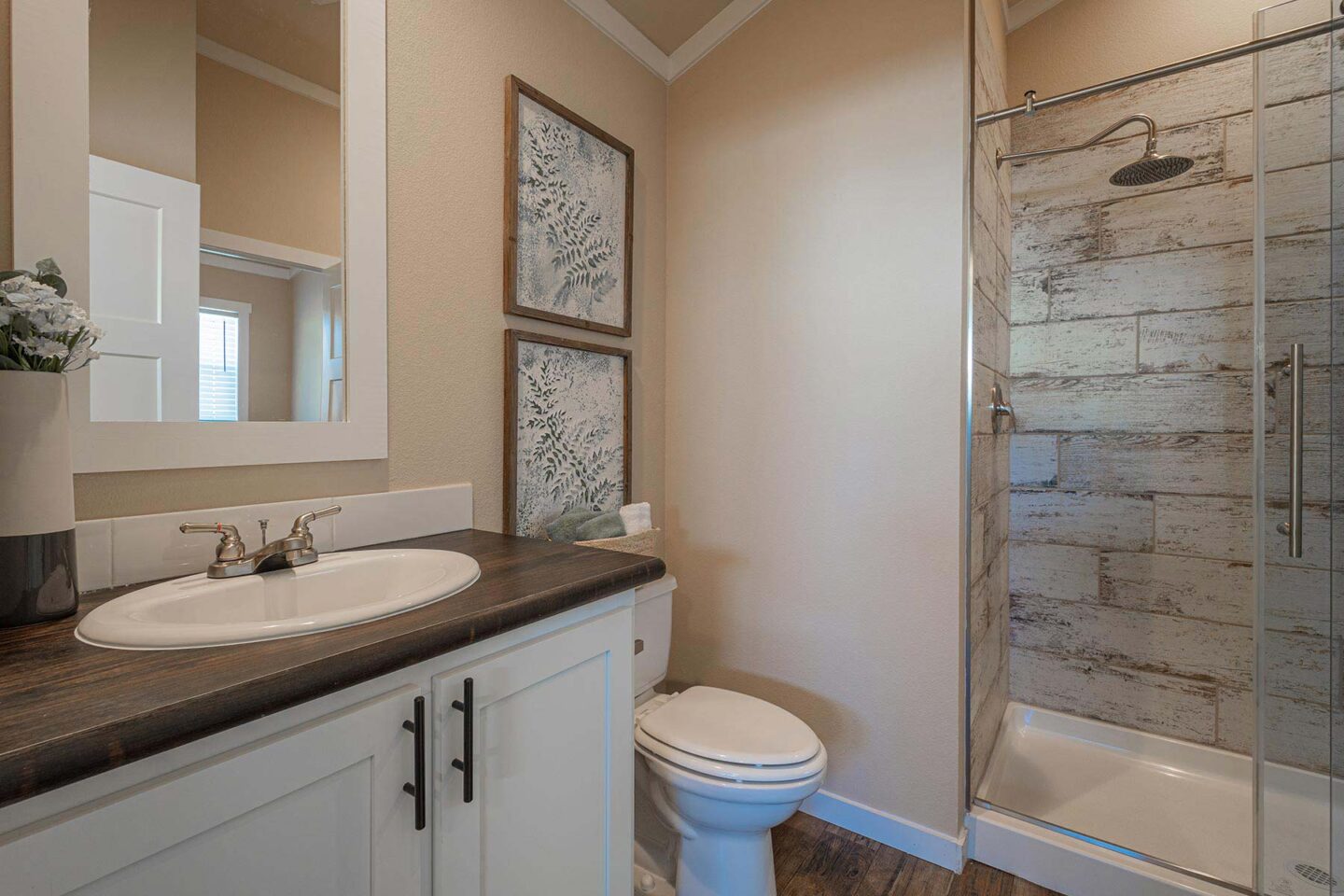 Cozy bathroom with wood accents, featuring a rustic shower with glass doors, white sink on a dark counter, and two botanical prints on the wall.