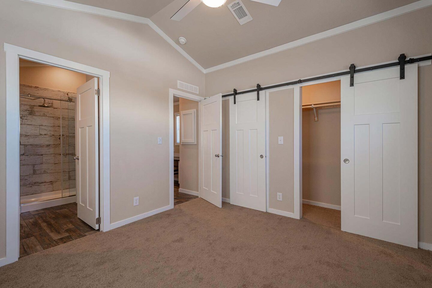 Empty, beige bedroom with carpet flooring, white trim, and ceiling fan. Features sliding barn doors revealing closets. Open door leads to rustic bathroom.