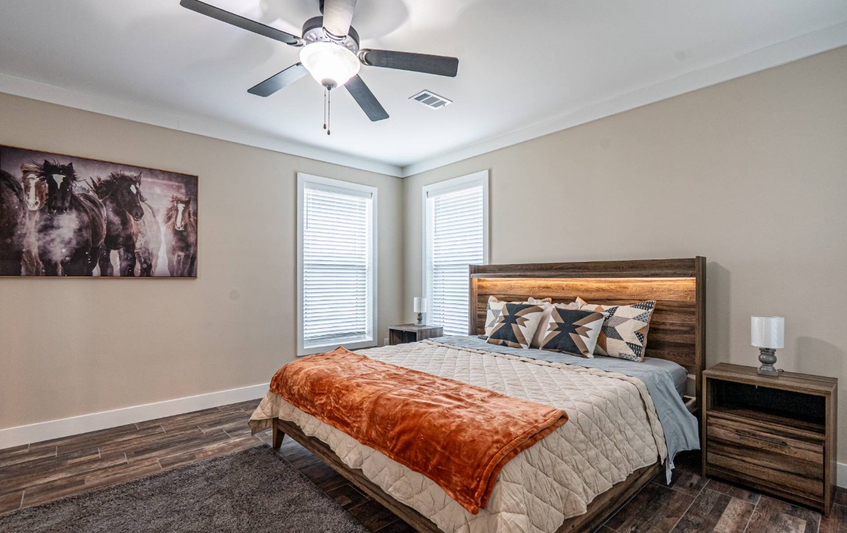 Modern bedroom with neutral tones, featuring a wooden bed, geometric pillows, and an orange throw. A ceiling fan and horse artwork add style.