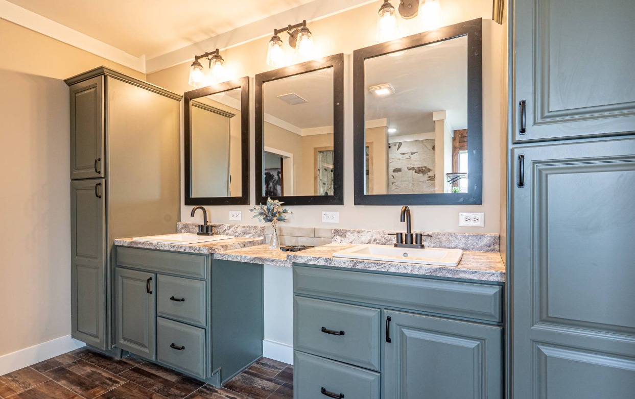 Modern bathroom with two green vanities, marble countertops, and black-framed mirrors. Soft lighting and wood floor create a warm, inviting tone.