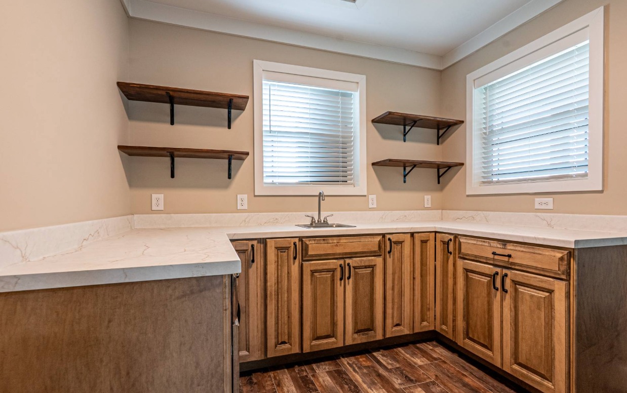 A cozy kitchen with wooden cabinets and floating shelves. White countertops and two windows with blinds create a bright, inviting atmosphere.