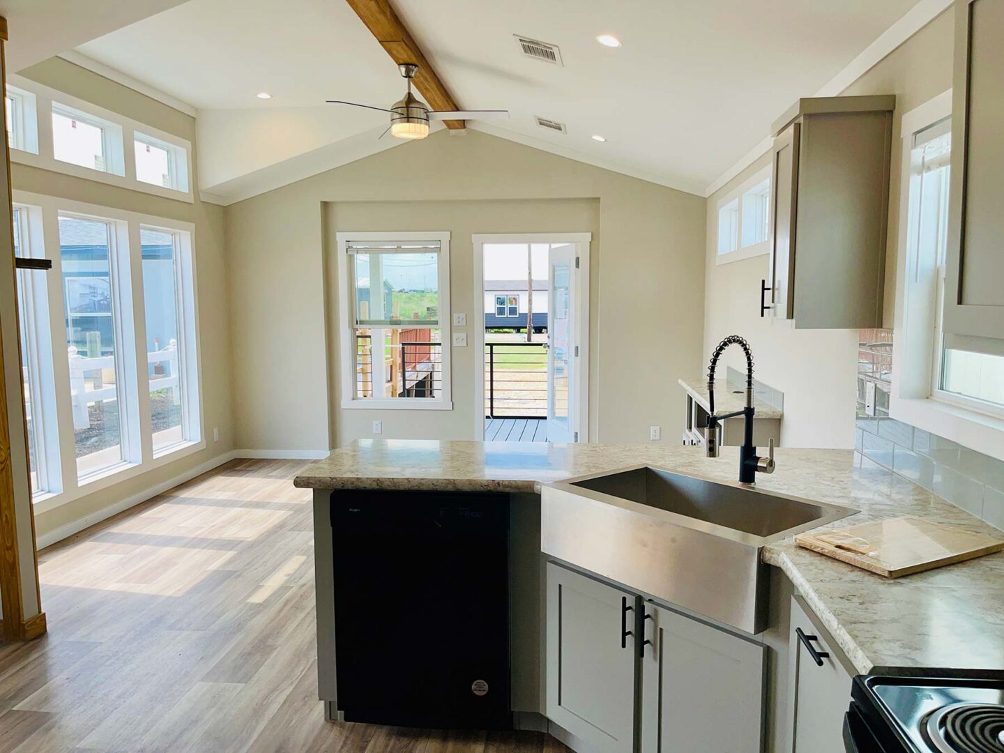 Bright modern kitchen with light wood floors, a high ceiling, stainless steel sink, and granite countertops. Large windows and a glass door let in ample sunlight.