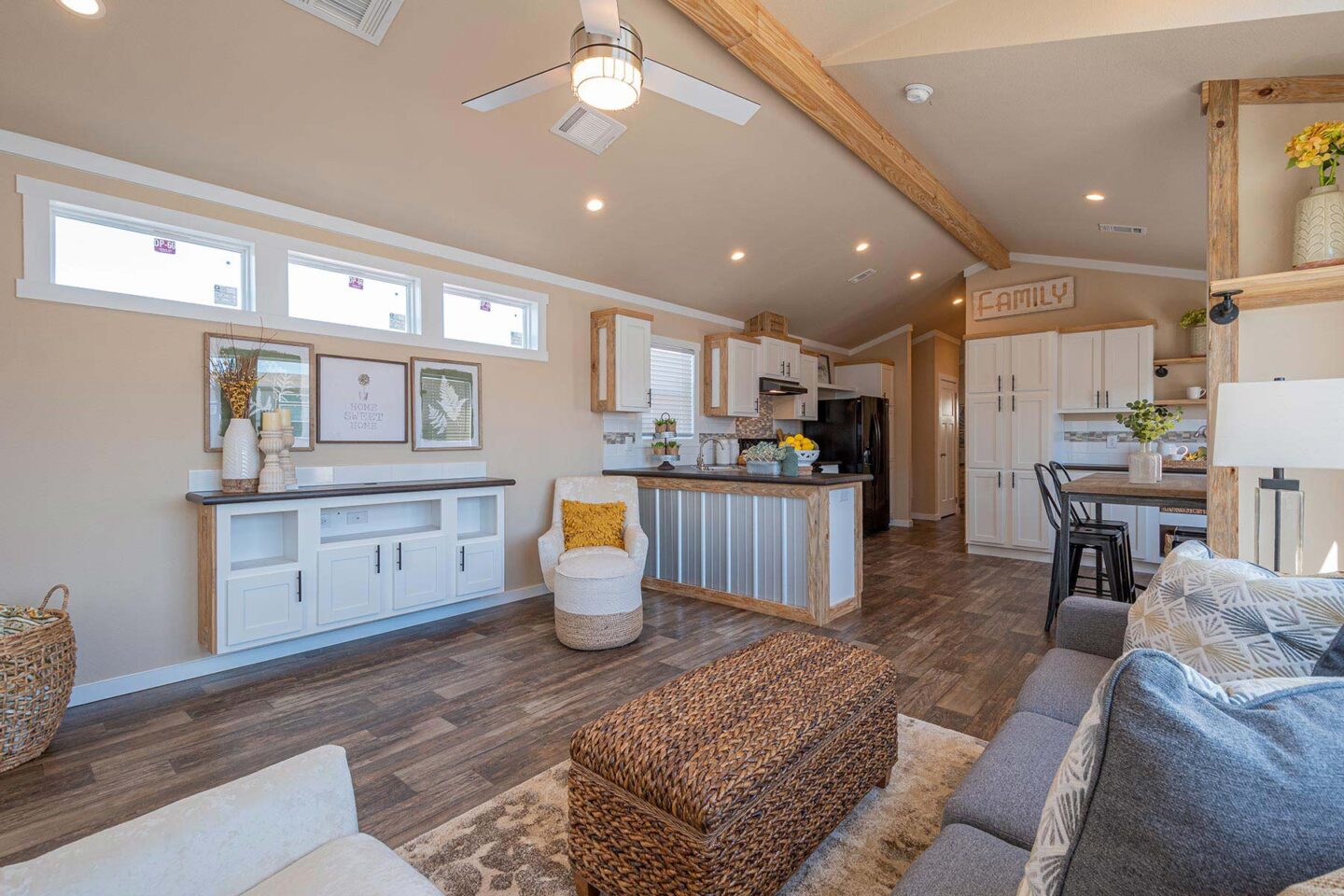 Cozy living room with a vaulted ceiling and wooden beams, featuring a wicker ottoman, rustic decor, and a kitchen island. Soft, warm tones create an inviting atmosphere.