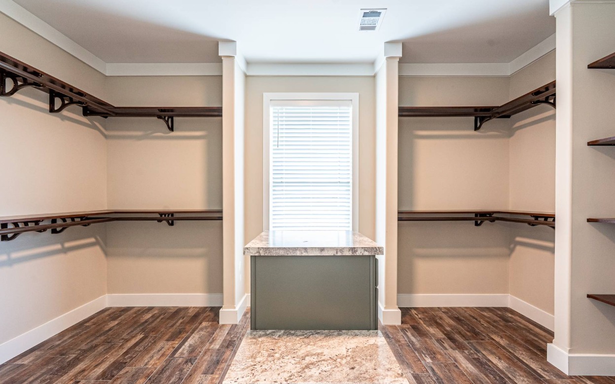 Spacious, empty walk-in closet with wooden shelves on beige walls, centered window with blinds, and a marble-top island on a rustic wooden floor.