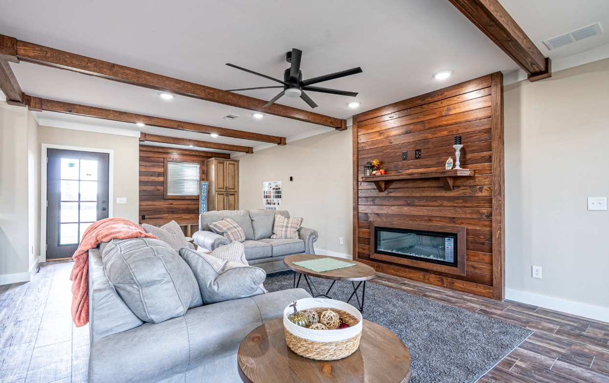 Cozy living room with wooden beams and a modern ceiling fan. Features gray sofas, decorative pillows, a warm wooden accent wall, and an electric fireplace.