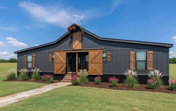 A modern barn-style house with dark siding, wooden shutters, and a gabled roof stands in a green lawn. Pink flowers and ornamental grasses line the path.