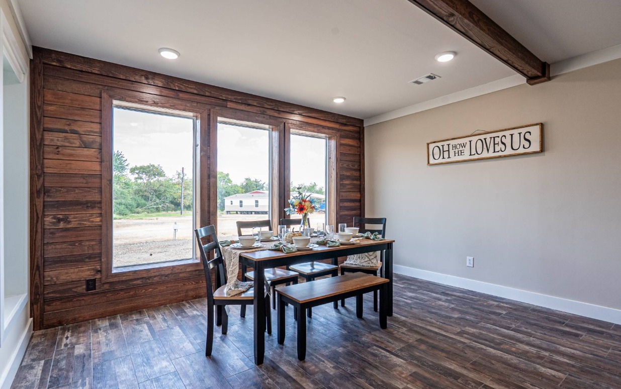 A rustic dining room with wooden floors and walls, featuring a set table with a bench and chairs. Large windows offer a view of greenery, and a sign on the wall reads "Oh how He loves us," adding a warm, homely feel.