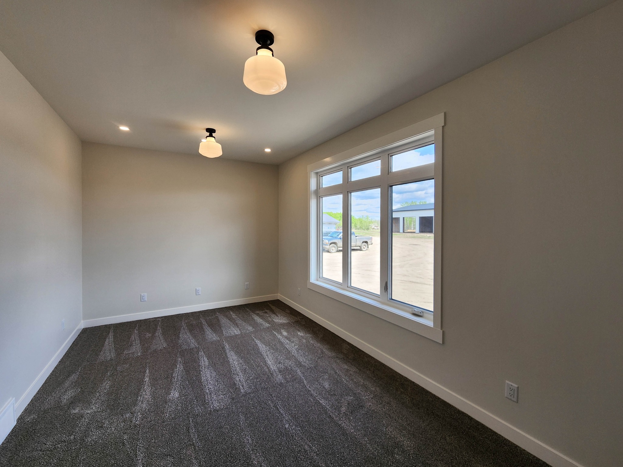 Empty room with beige walls, two ceiling lights, and a large window showing an outdoor view. Gray carpet covers the floor, creating a clean, tranquil space.