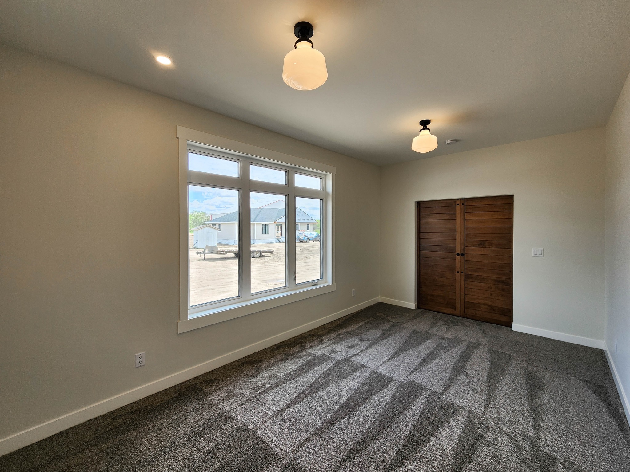 A bright, empty room with two ceiling lights and a large window overlooking a construction site. It features beige walls, gray carpet, and a wooden closet door.