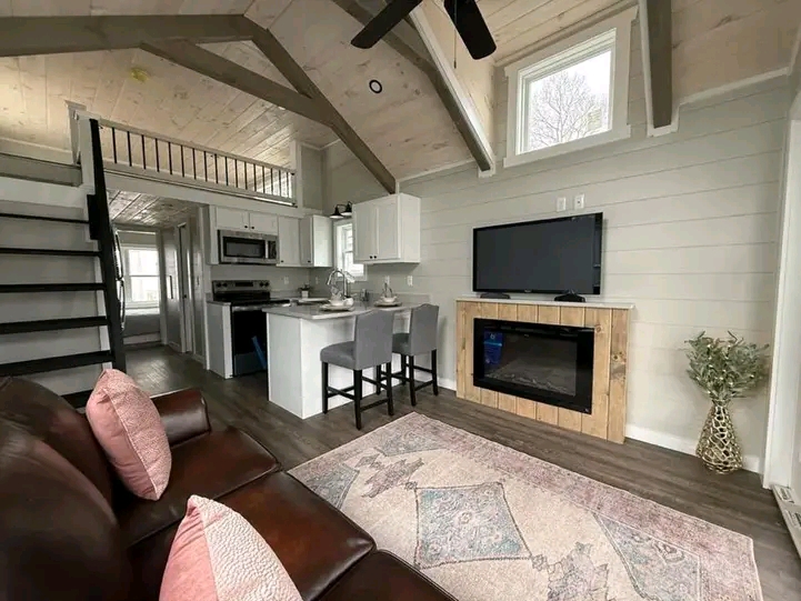 Cozy living area with vaulted ceiling, featuring a brown leather sofa with pink pillows, a flat-screen TV on a wooden stand, and a small dining island with gray chairs near the kitchen. A loft with black railing and wooden stairs is visible. The space has light gray walls and warm wood floors, creating a welcoming feel.