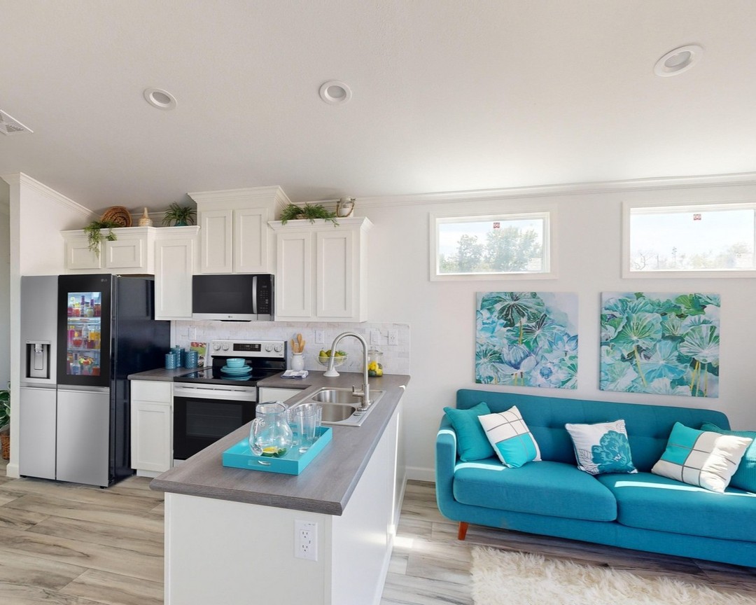 Modern kitchen and living area with white cabinets, stainless steel appliances, and a gray countertop. A vibrant teal couch sits nearby, adorned with white and teal pillows. The walls are decorated with colorful abstract artwork, creating a fresh and inviting atmosphere.