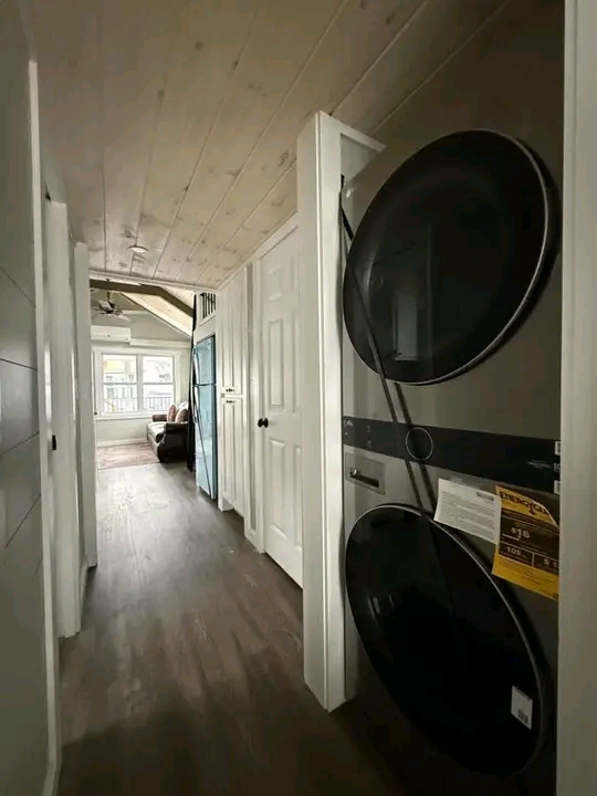 A narrow hallway with wooden flooring leads to a bright living room. On the right, a stacked washer and dryer sit in a closet. Warm and cozy atmosphere.