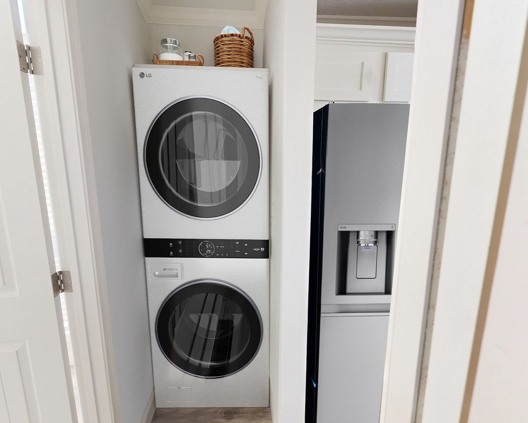 Stacked washer and dryer with digital display in a narrow laundry nook, adjacent to a silver fridge. Basket and jar on top add a cozy touch.