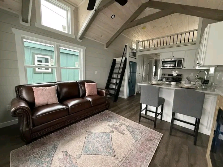 Cozy tiny home interior with vaulted ceiling. Leather sofa with pink cushions, loft with ladder, kitchen island with two stools, and large windows.