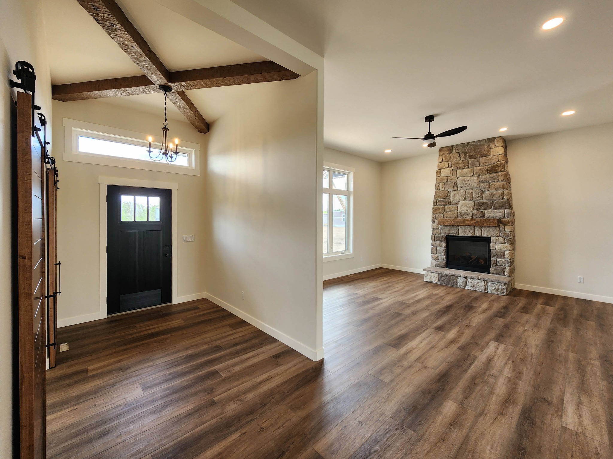 Spacious living area with wood floors, a stone fireplace, ceiling fan, and large window. Entryway with dark door and wooden beams overhead. Cozy and inviting atmosphere.
