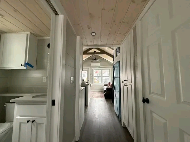 Narrow hallway of a cozy, modern tiny home with light wood ceiling and white cabinets. Bright natural light from large windows creates an inviting atmosphere.