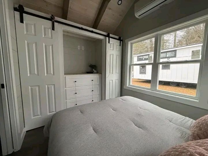 Cozy bedroom featuring a compact closet with sliding doors and a white dresser. Large window offers a view of parked trailers outside, under a vaulted ceiling.