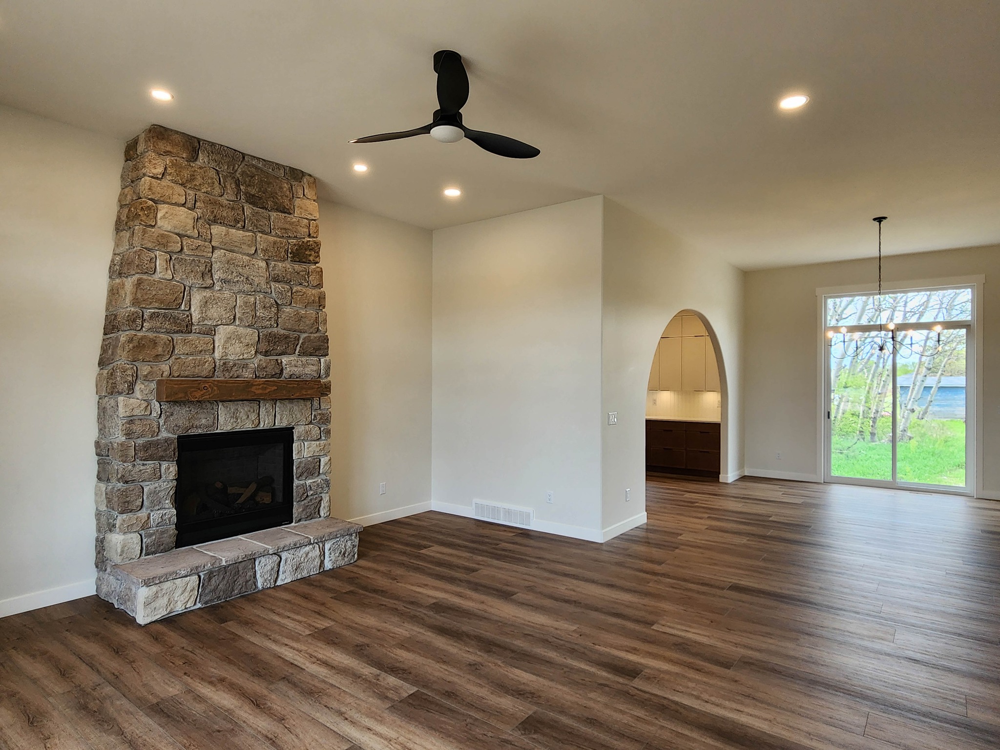 Spacious living room with wood flooring and a stone fireplace. Features modern ceiling fan and large window, creating a cozy yet airy atmosphere.