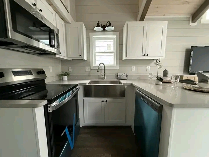 Modern tiny house interior with a vaulted ceiling, featuring a compact kitchen with white cabinets, a breakfast bar, and dark wood floors. A loft area and black ladder enhance the cozy yet elegant ambiance.
